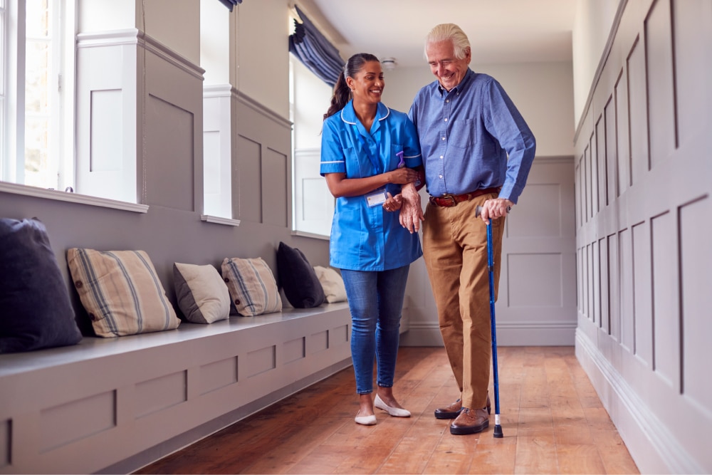 An elderly man is walking down a care home corridor with a nurse holding his arm. They are both smiling.