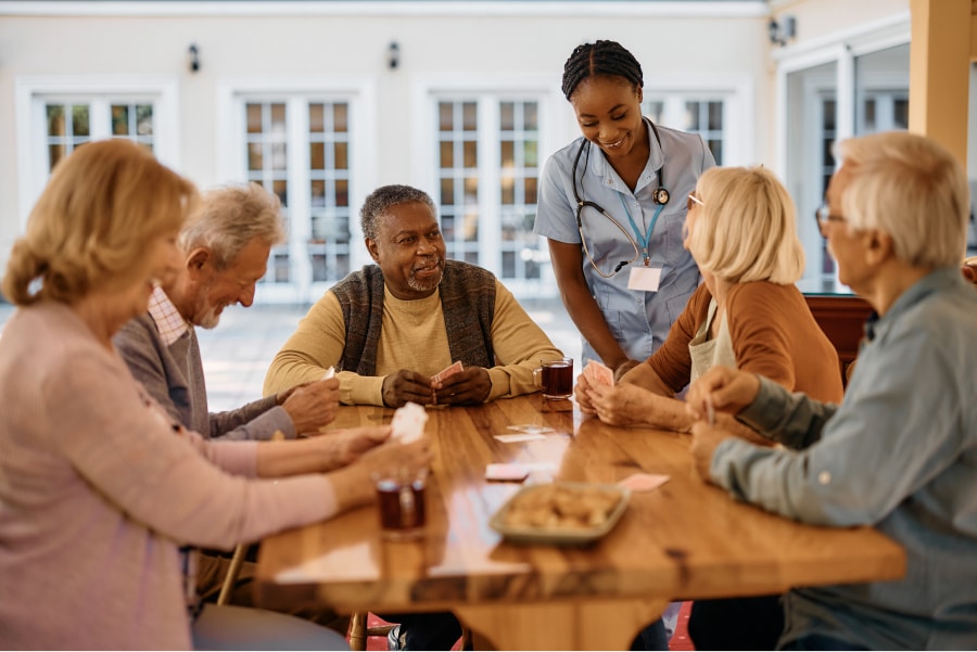 A group of seniors playing cards around a wooden table, with a care home nurse leaning over them and smiling.