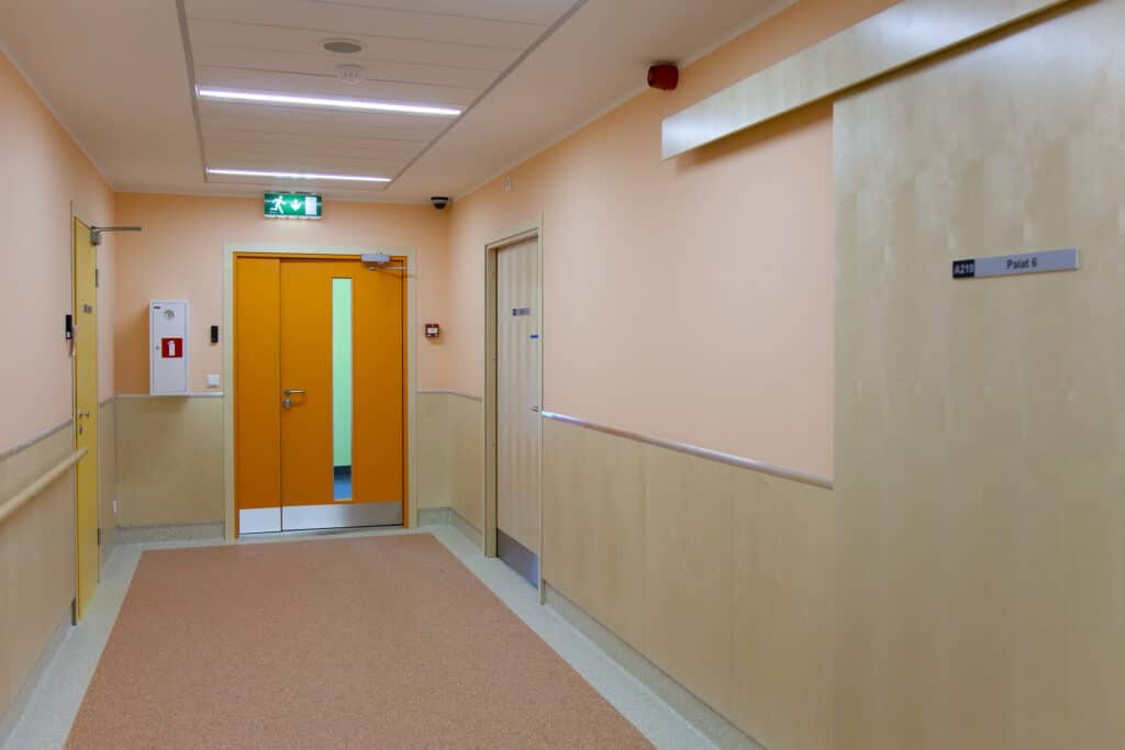 A fire door at the bottom of a hospital corridor. You can see a Fire Exit sign above it and a fire alarm trigger next to it.