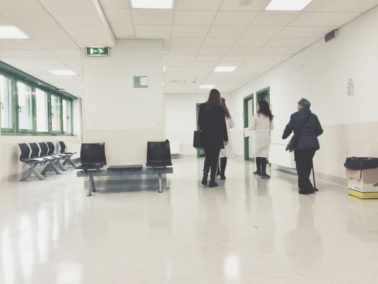 Wide view of a bright hospital corridor with staff and a visitor walking between clinical rooms, seating, emergency exit signage and fire door openings, representing a live healthcare environment where fire compartmentation, safe escape routes and ongoing compliance management are critical